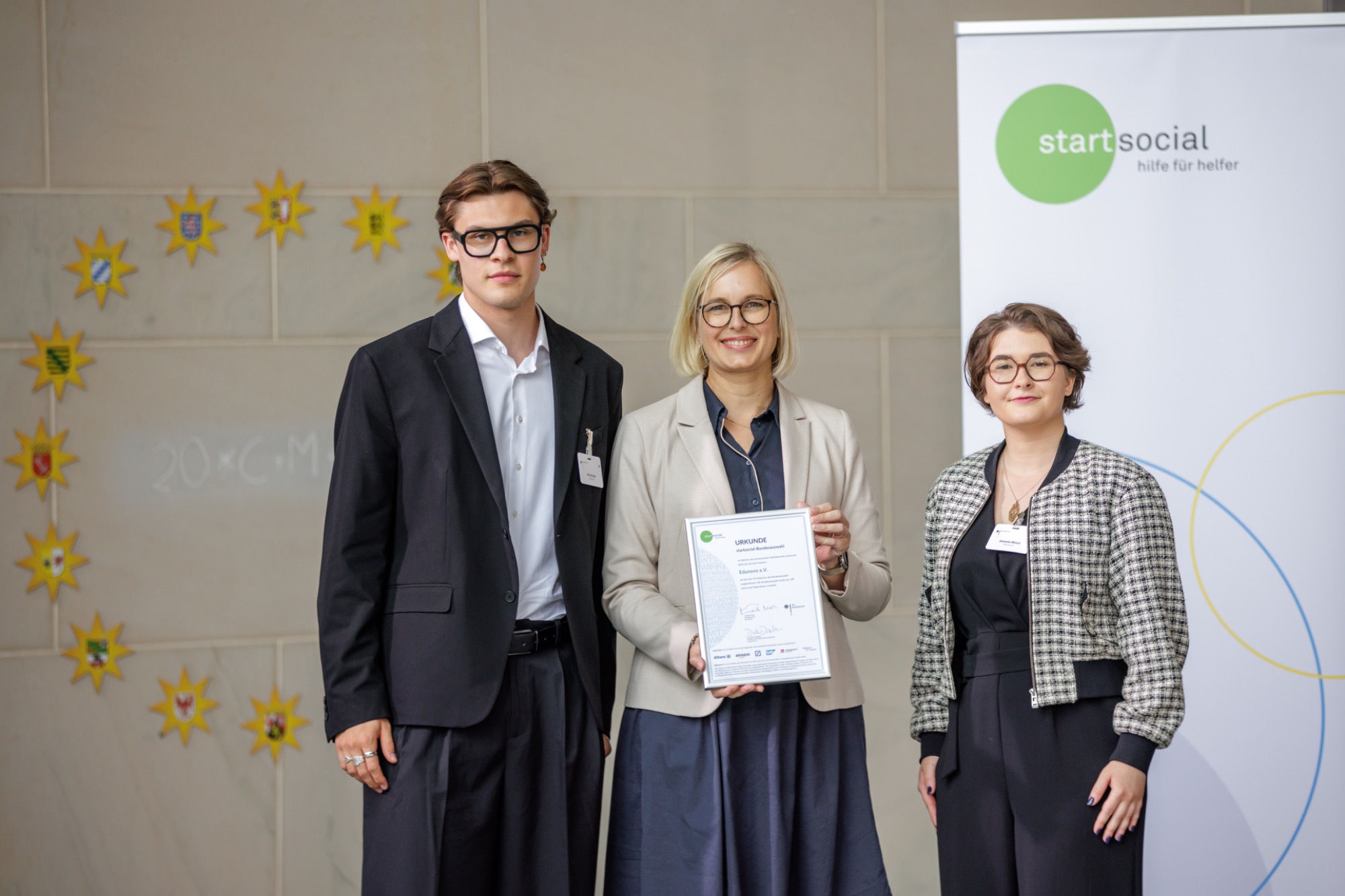 Edunovo in der startsocial-Bundesauswahl 2026: Nils und Emmelie treffen Staatsministerin Dr. Christiane Schenderlein im Bundeskanzleramt Drei Personen stehen zusammen, halten ein Zertifikat, im Hintergrund steht ein Banner.
