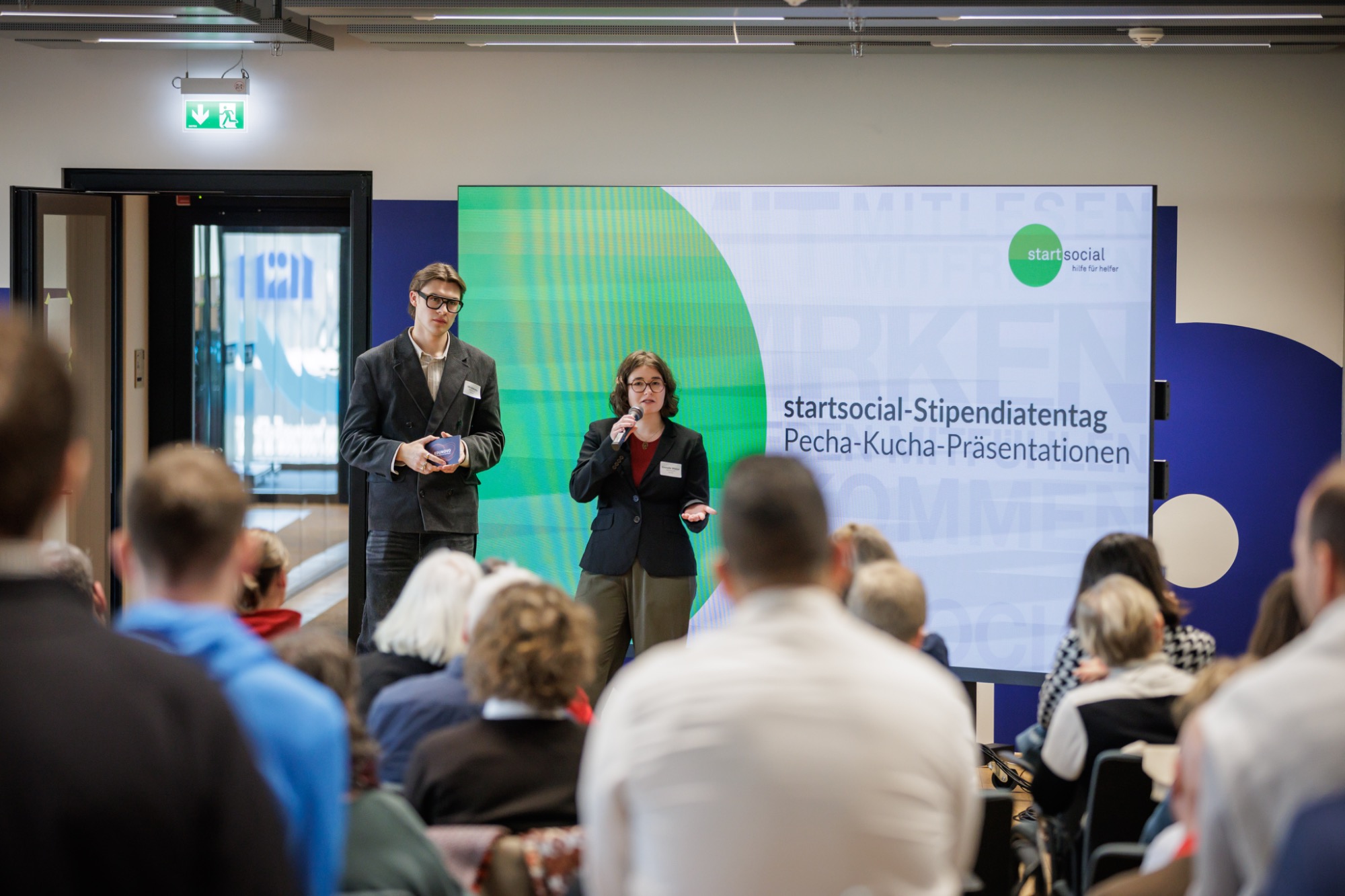 Edunovo beim startsocial-Stipendiatentag in Berlin: Nils und Emmelie stellen unsere Arbeit für mehr Bildungsgerechtigkeit vor Zwei Personen präsentieren auf einer Bühne vor Publikum, im Hintergrund ein großes Display.